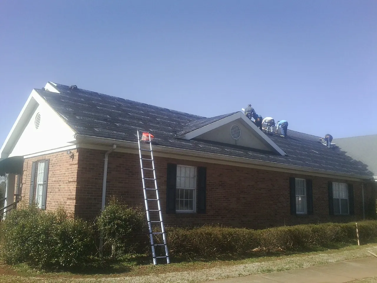 Crew tearing shingles off a brick ranch roof with a ladder set — Emergency Roof Repair work by our crew in Ennis